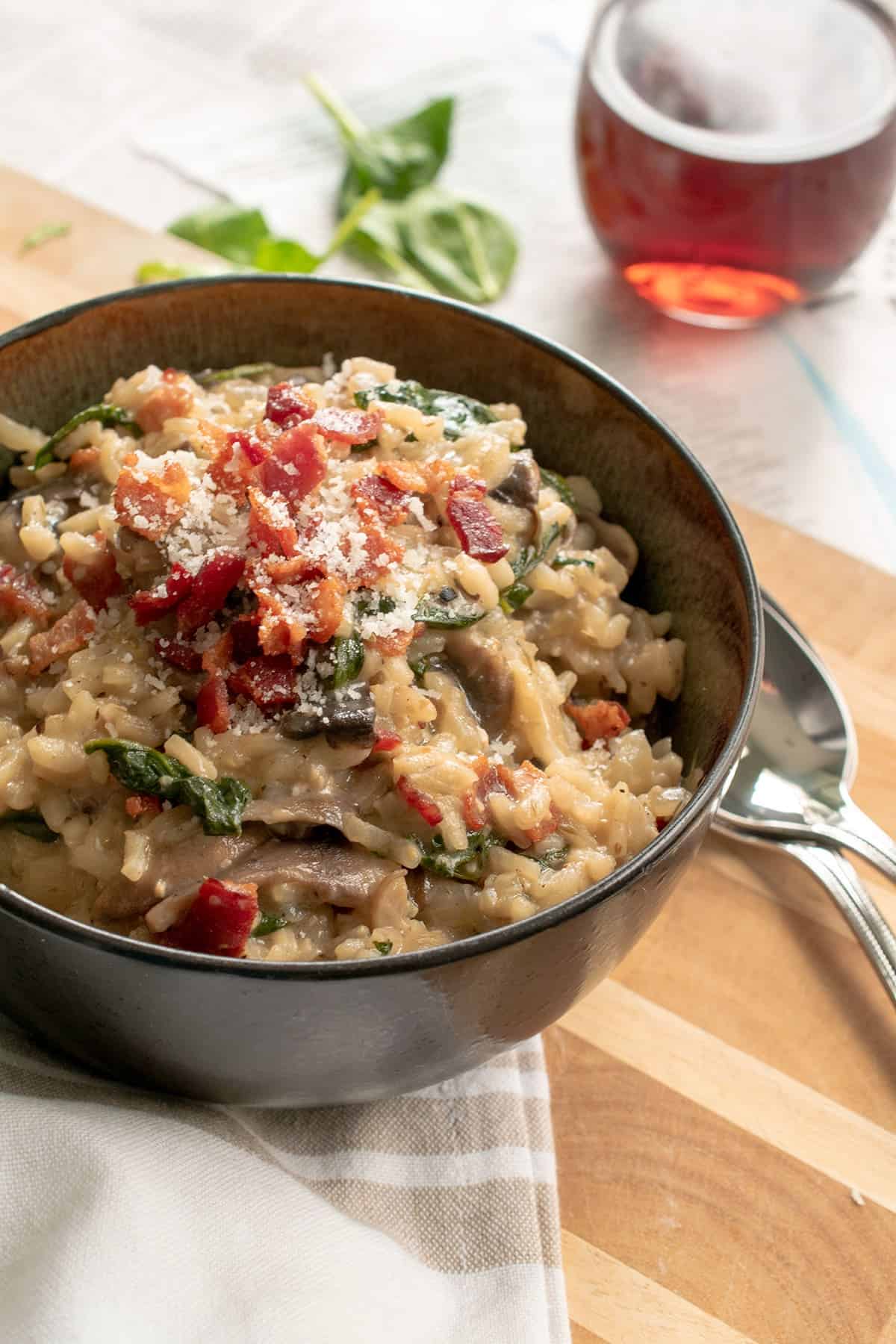 A bowl of bacon mushroom risotto on a wood surface with metal spoons next to it.