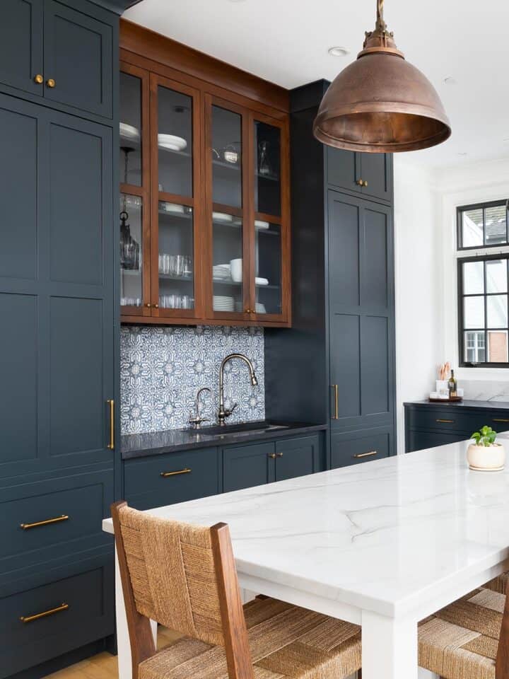Behr Midnight blue painted cabinets in a classic old world kitchen with modern white accents and backsplash. Wicker chairs, copper light fixtures, and wood floors add warmth.