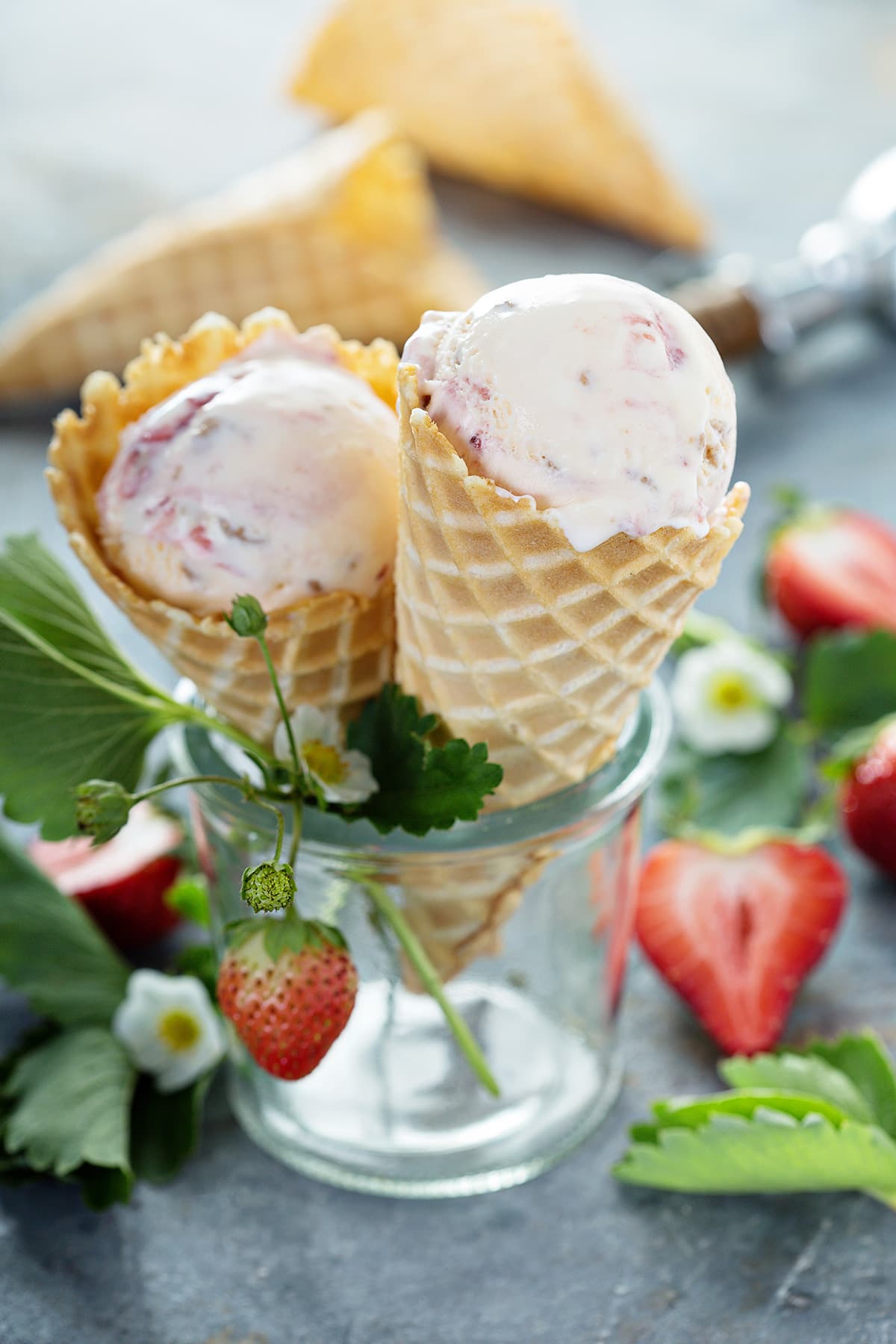 Two waffle cones with strawberry cheesecake ice cream in glass with vine of strawberries next to waffle cones and ice cream scoop.