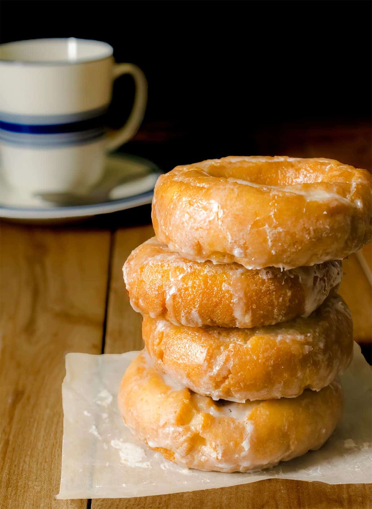 Stack of classic glazed sour cream cake doughnuts on parchment paper with cup of coffee on the side