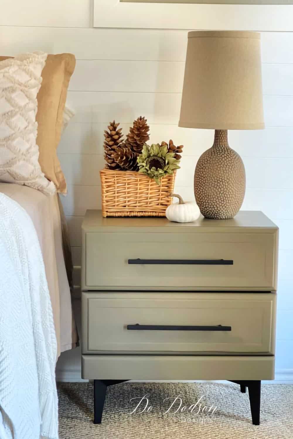 Olive green nightstand with black accents with basket with pinecones next to lamp.