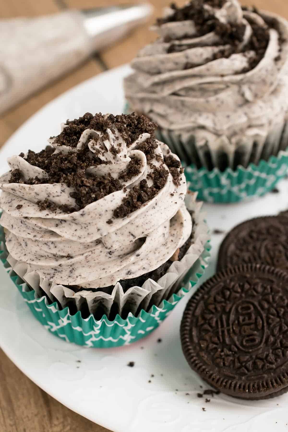 Two chocolate cupcakes in turquoise liners on white plate next to stacked Oreo cookie.