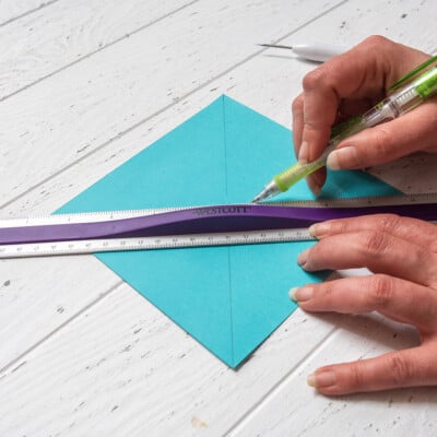 Colorful craft paper marked by hands with pencil and metal ruler on white surface.