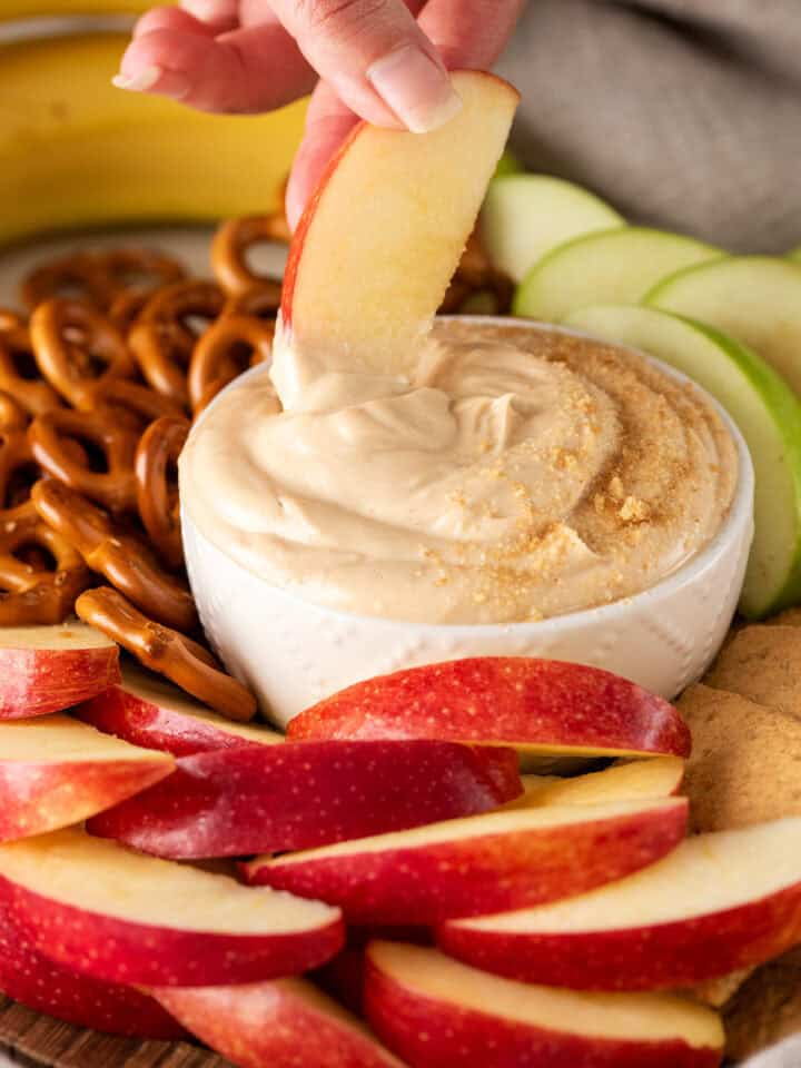 A hand dipping an apple slice into a bowl of sweet peanut butter dip.