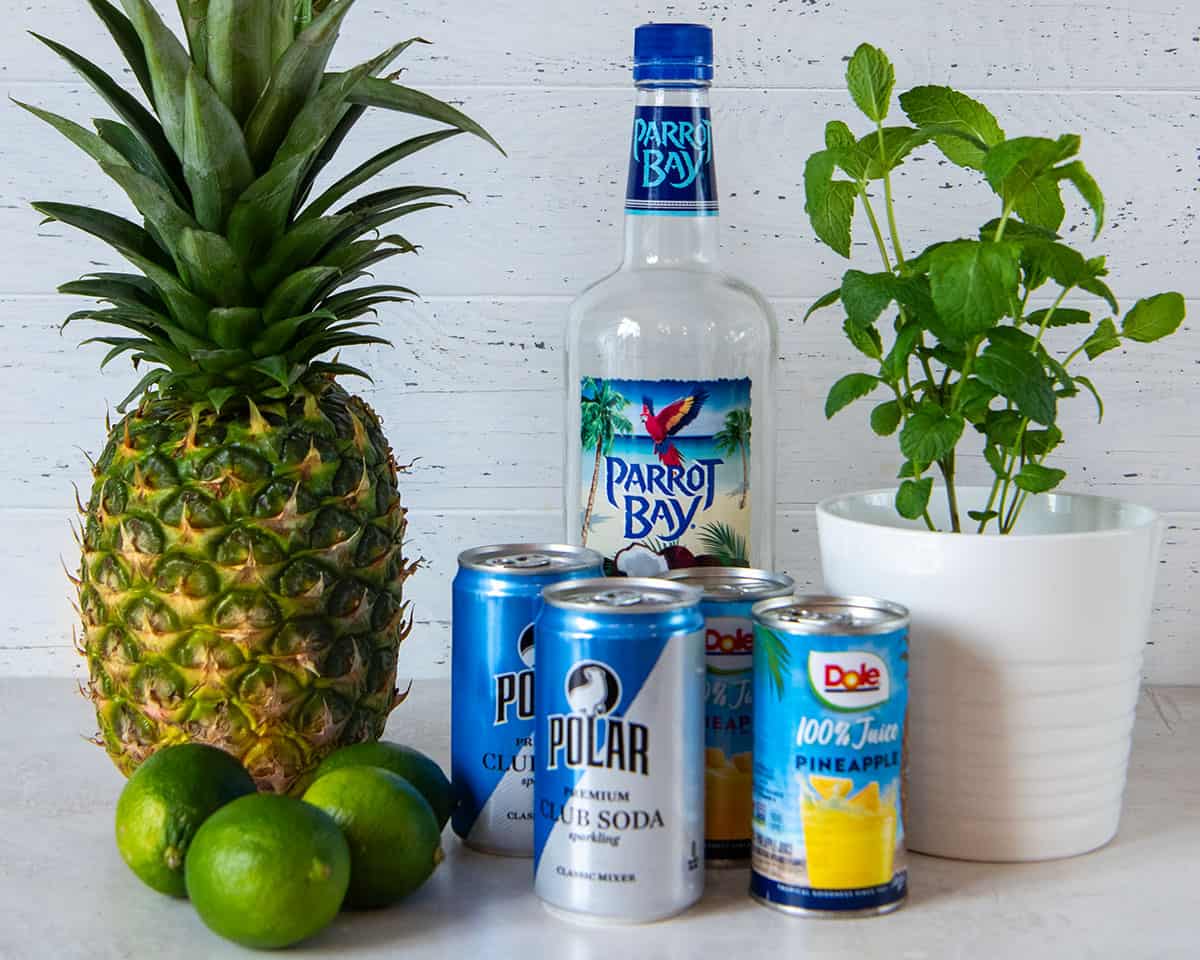 Ingredients to make a Pineapple mojito with coconut rum, mint, pineapple juice, and club soda arranged on a table.