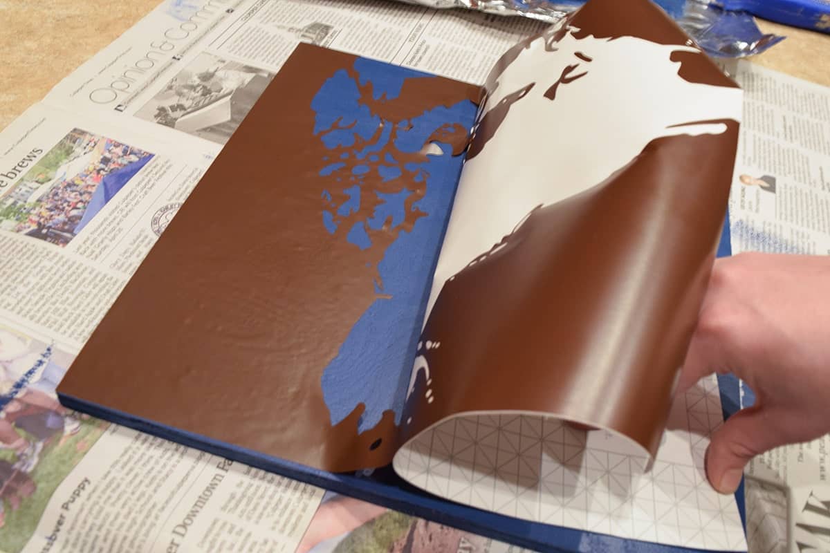 Peeled away vinyl stencil of world map section applied to blue wooden square over newspaper.