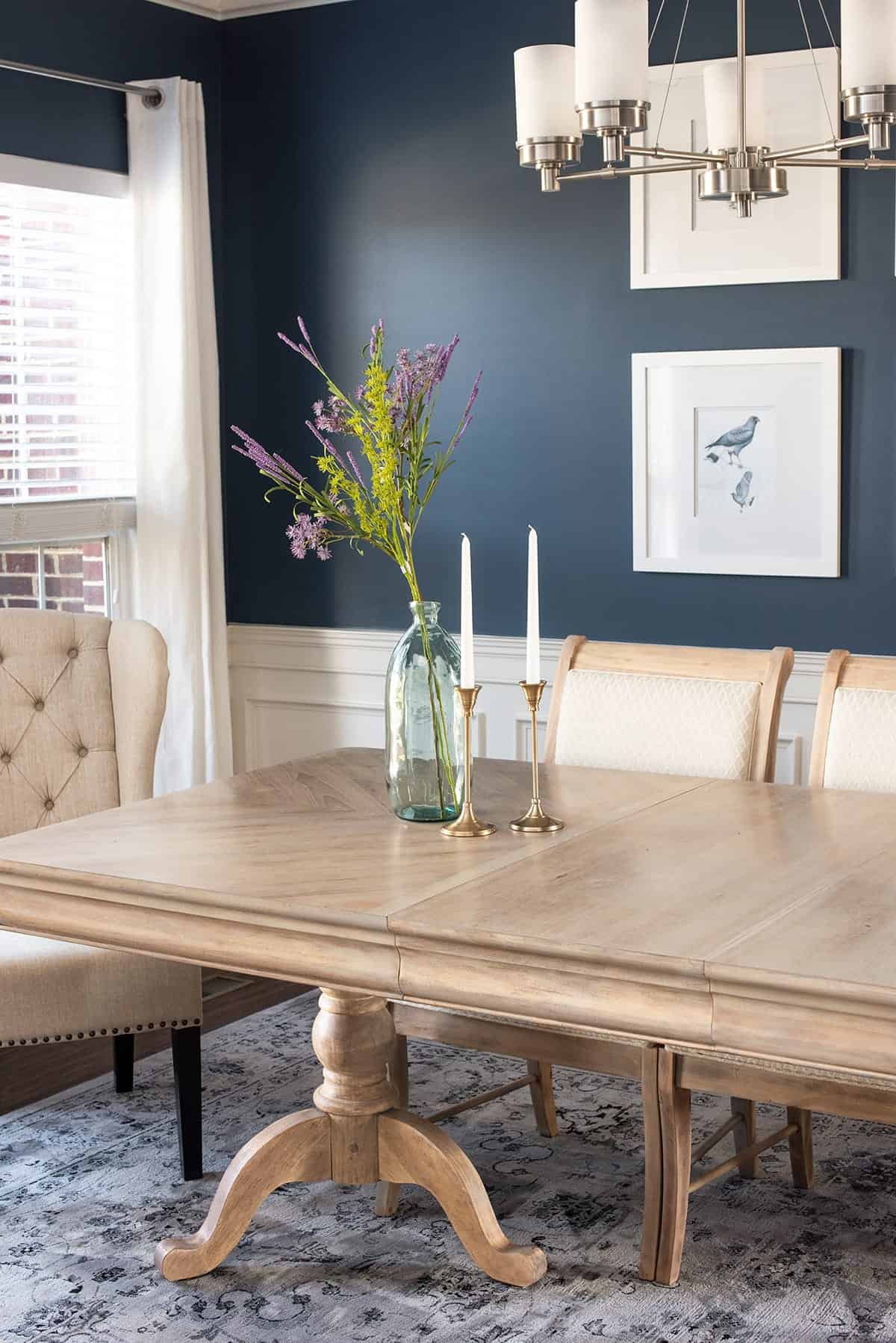 Finished natural raw wood dining room table and dining room chairs. Cerulean blue walls with bird print, bud vase and brass candlesticks adorn table.
