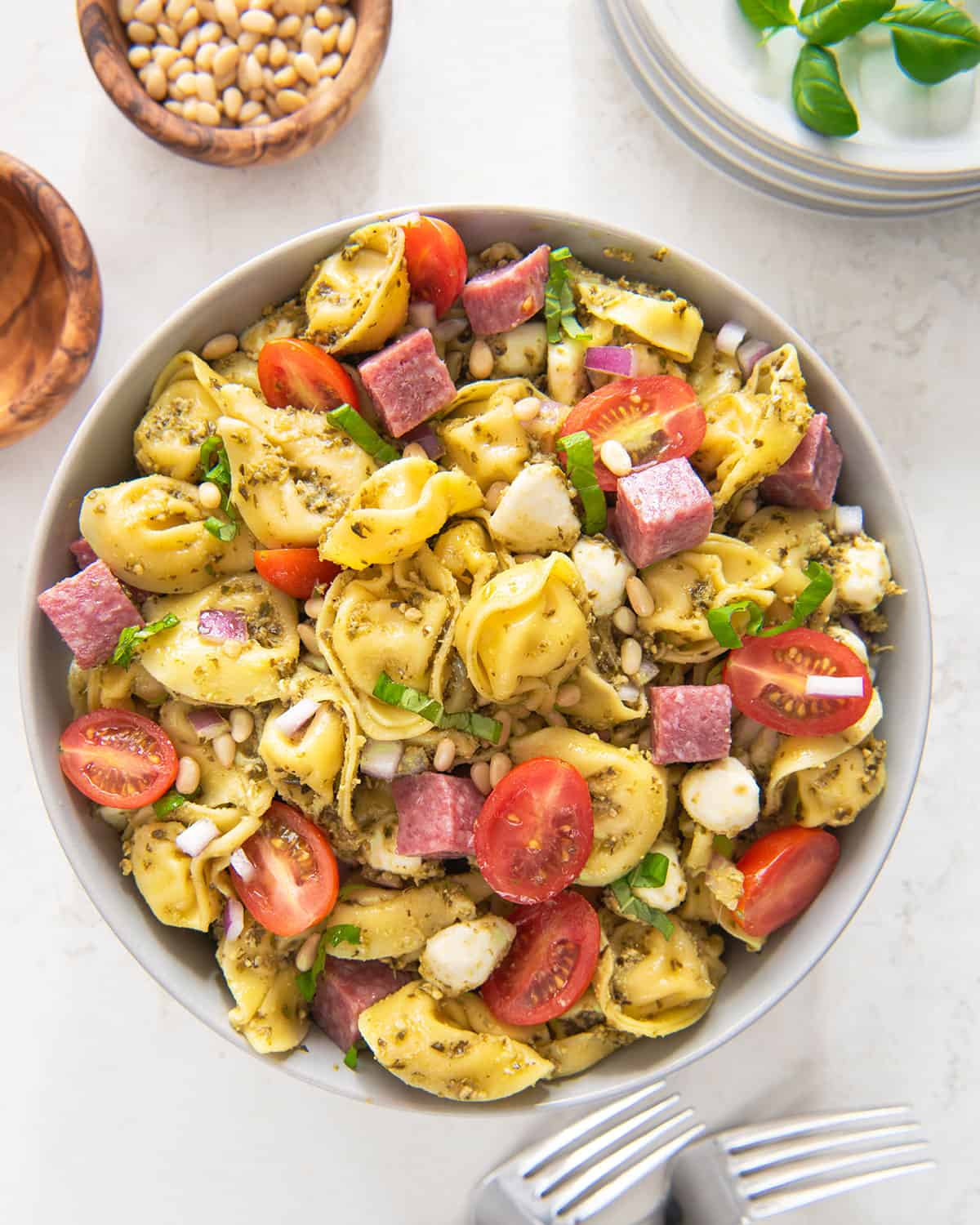 Top-down view of a bowl of pesto tortellini pasta salad with salami, tomatoes, pine nuts, and basil surrounded by ramekins and forks.