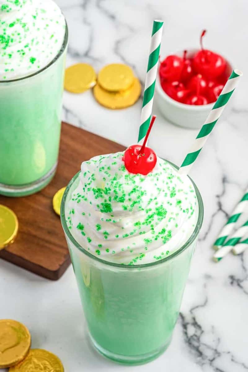 Shamrock shake with whip cream and green sprinkles topped with a maraschino cherry and green striped straws.