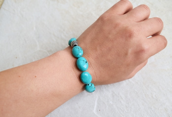 Woman's hand with simple turquoise DIY beaded bracelet over white marbled surface.