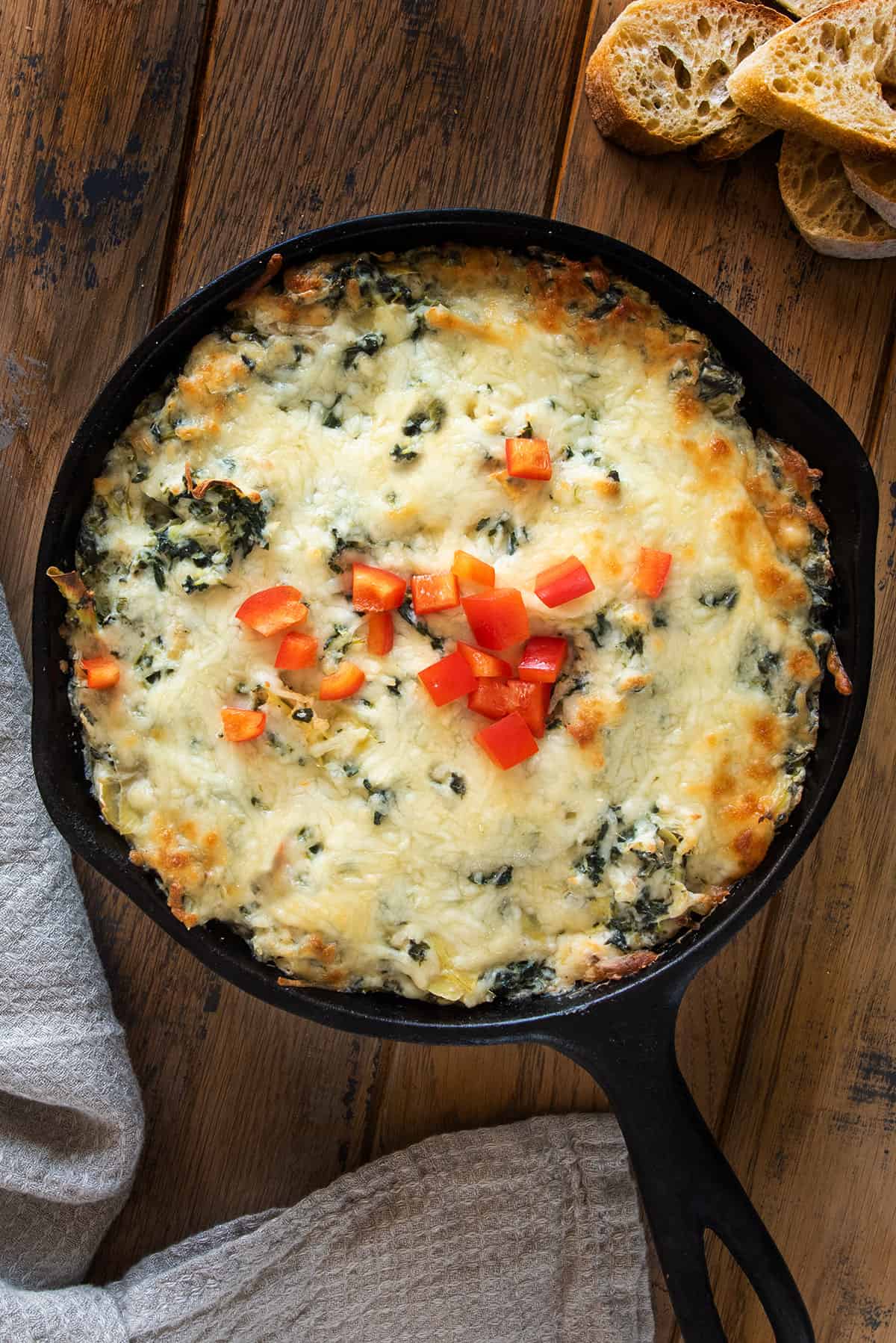 Overhead of baked spinach artichoke dip with red peppers on top in a cast iron pan on a wood slab.