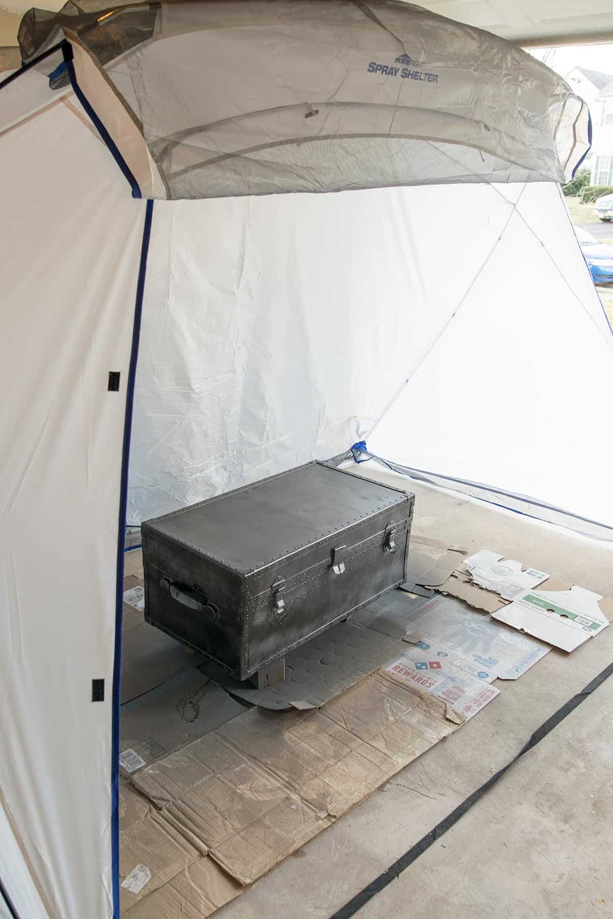 Painting an antique metal steamer trunk with spray paint. Trunk is located inside a spray shelter.