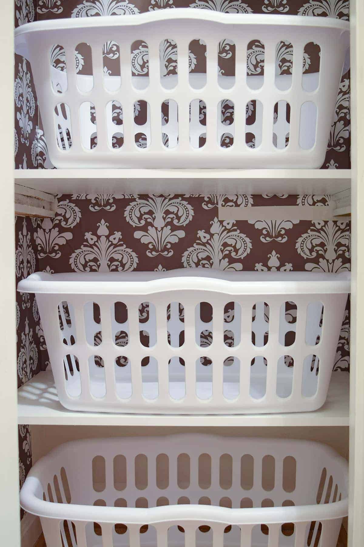 Inside of laundry closet with peel and stick brown and white wall covering, white shelves, and 3 white laundry baskets.