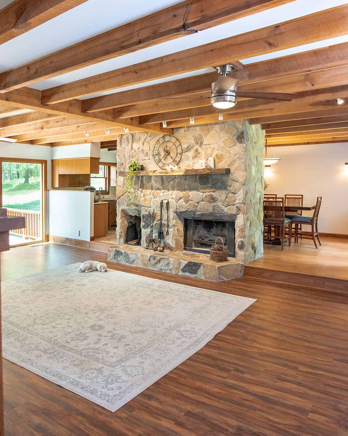 Rustic cabin living room with wood floors and wood beam ceilings, while a giant stone fireplace stands in the middle.