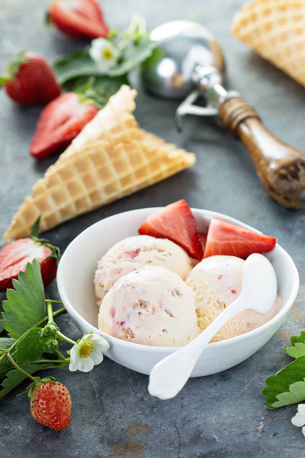 Strawberry cheesecake ice cream in white bowl with sliced strawberries and white spoon, on granite countertop.