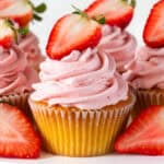 A strawberry frosted vanilla cupcake with a strawberry halve on top and flanked by more cupcakes.