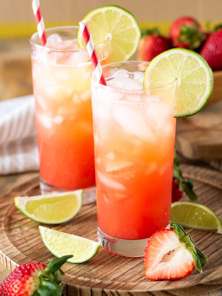 Two glasses of strawberry limeade with lime garnish on a wood background.