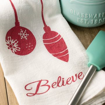 Christmas Stenciled Tea Towels laid on a table jumping reindeer, ornaments, Christmas Tree silhouettes, and text.