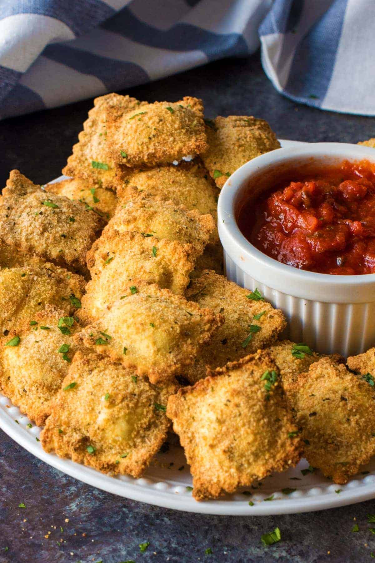 Toasted Ravioli on a white plate with marinara in white cruet. White and blue dish towel in the background.