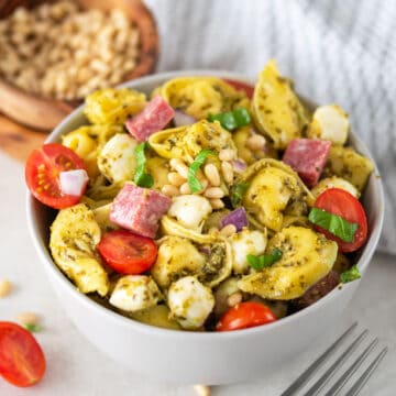 A bowl of pesto tortellini pasta served cold with mozzarella, salami, and tomatoes on top.