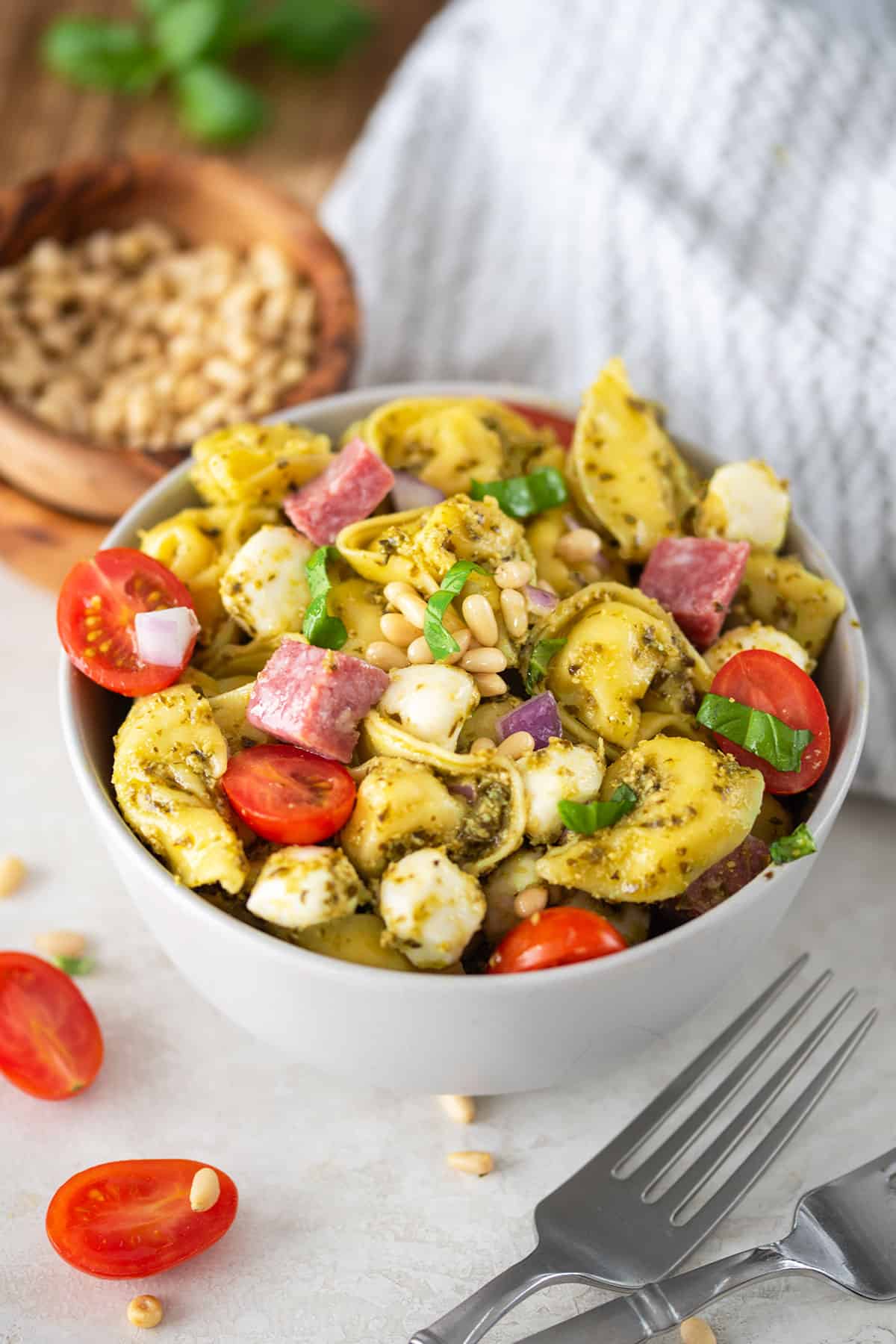 A bowl of pesto tortellini pasta served cold with mozzarella, salami, and tomatoes on top.