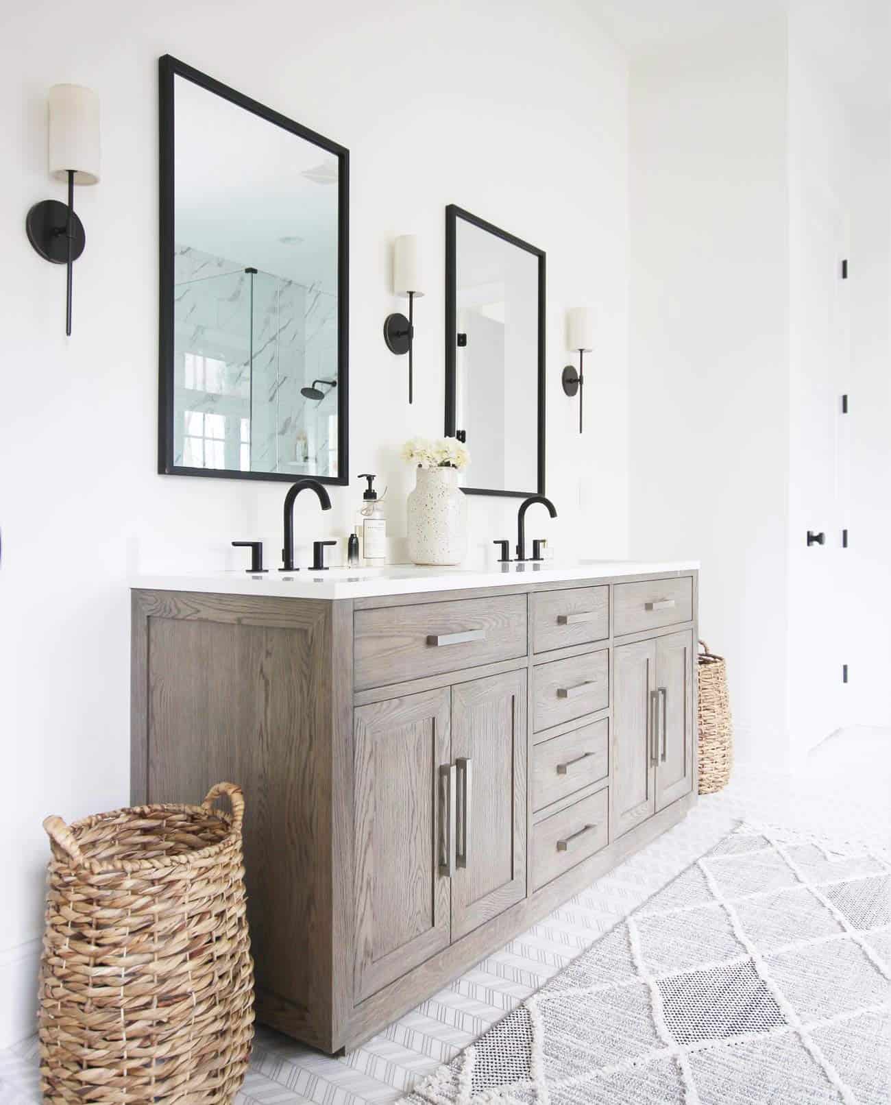 Transitional bathroom with grey wood vanity and black accents. Sconces on either side of the mirror and natural baskets on the floor.