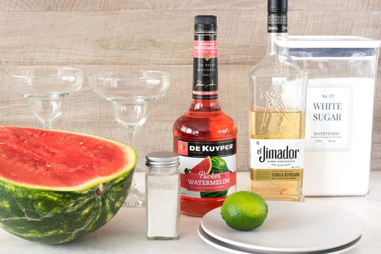 Watermelon Margarita ingredients laid out on a table including tequila, sugar, lime, watermelon, salt, and watermelon pucker.