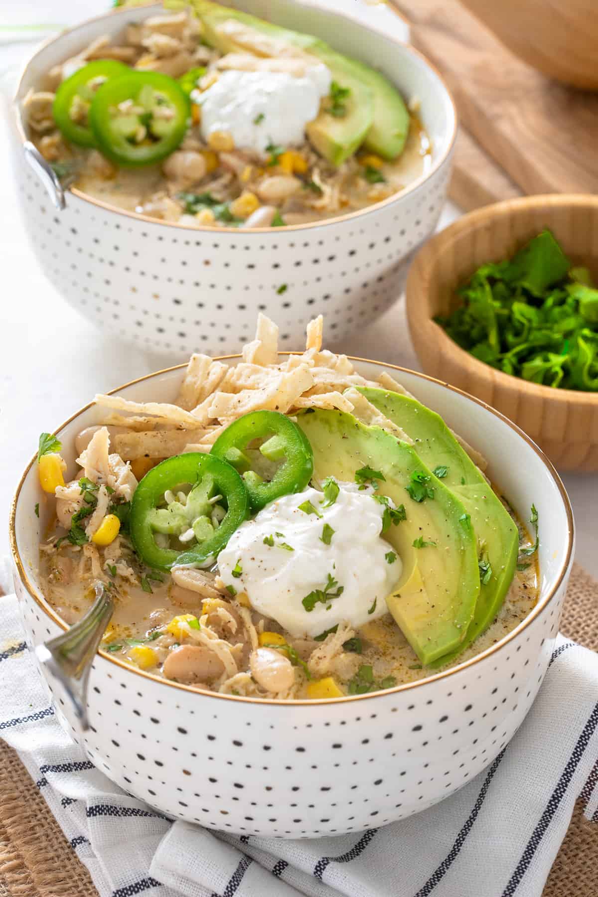 Two Bowls of white chicken chili on white background garnished with tortilla strips, jalapeno, avocado, and cilantro.