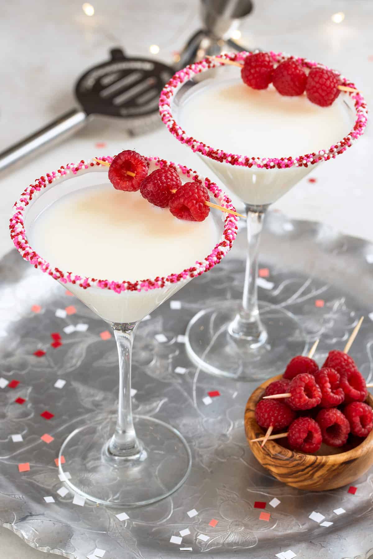 Two White Chocolate martinis with raspberry garnish on a silver tray for Valentine's day with a bowl of garnishes on the side and confetti.
