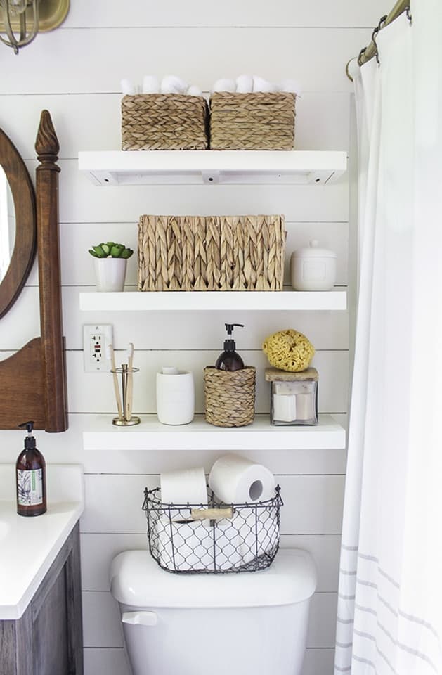Three white floating shelves above commode with wicker storage baskets, toiletries, and small linens.