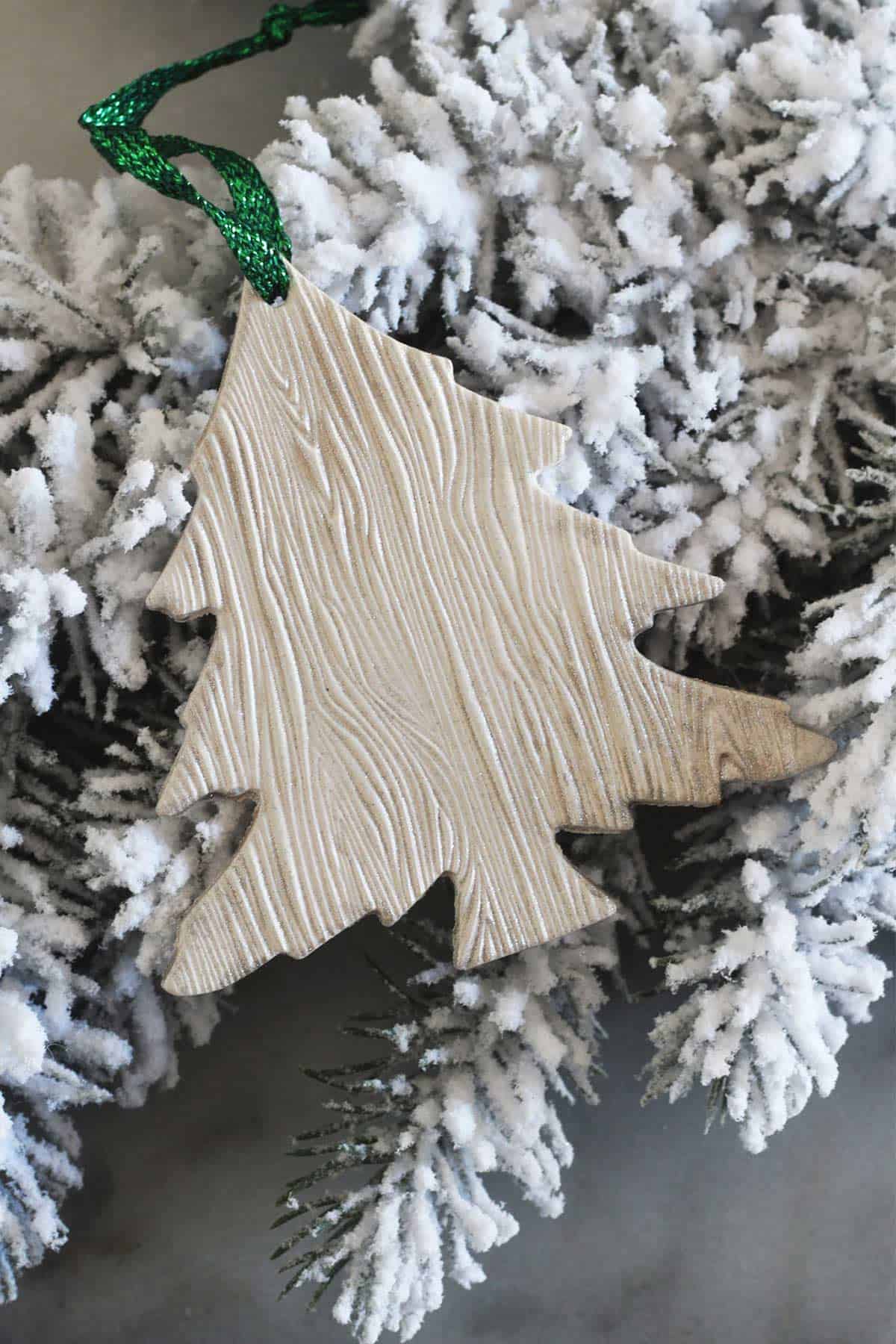 Clay Christmas tree shaped Christmas ornament with wood grain style pattern in natural neutral pattern hung from frosted branch with sparkly green ribbon.
