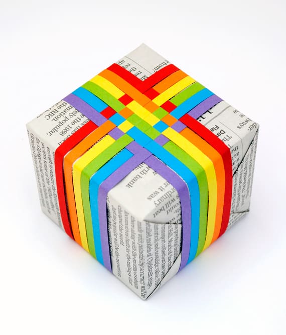Gift covered in newspaper, topped with rainbow colored paper basket weave.