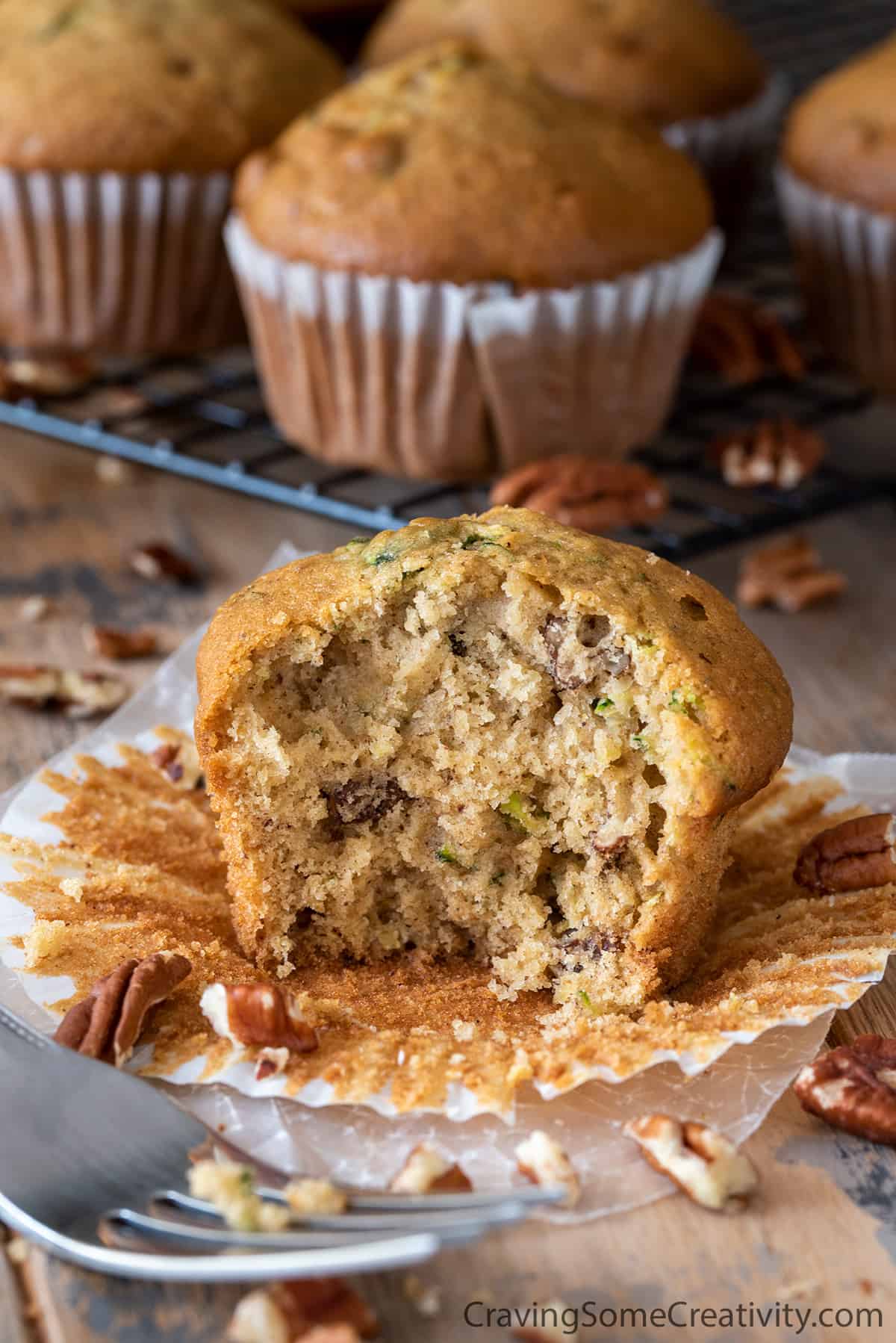 Zucchini Muffin with bite taken out to show center texture on a wood background with whole muffins.